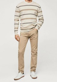 Hombre vistiendo pantalones beige, un suéter de rayas blancas de manga larga, zapatillas blancas, de pie con una mano en el bolsillo sobre un fondo liso.