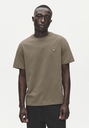 Jeune homme noir portant un T-shirt marron uni à manches courtes avec un petit logo rond sur la poitrine, debout devant un fond blanc.