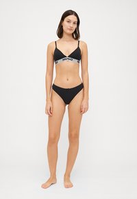 Μαύρο μπικίνι με τριγωνική μπλούζα και αντίστοιχες κιλότες, διαθέτει φαρδύ ελαστικό ύφασμα με λογότυπο και γκρι κείμενο. Απαλή, λεία υφή.