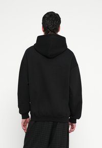 Čierna mikina s veľkou, nastaviteľnou kapucňou, spustenými ramenami a rebrovanými manžetami a lemom. Materiál vyzerá mäkko a hladko.