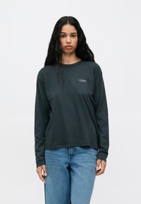 Ciemnozielona koszula z długim rękawem i okrągłym dekoltem, z małym białym logo "OBEY" na lewej piersi, połączona z niebieskimi dżinsami.