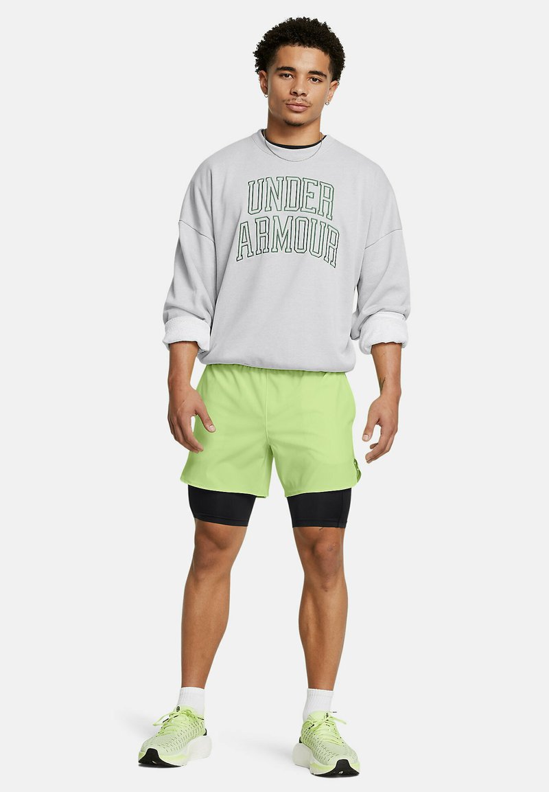 Hombre que lleva un sudadera de "Under Armour" gris claro, pantalones cortos neón verde sobre pantalones cortos de compresión negros y zapatos deportivos a juego en neón verde.