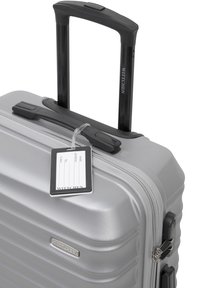 Silver hard-shell suitcase with a textured finish, retractable black handle, and clear luggage tag attached. Features a zippered closure.