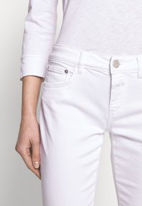 Jeans de mezclilla blanca con un diseño ajustado. Presenta un bolsillo frontal, cierre de botón de metal y detalles de costura sutiles. La textura parece suave.