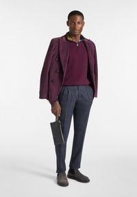 Bordeauxfarbener Cord-Blazer, tiefviolette Zip-Jacke, gestreifte navyfarbene Hose, graue Wildleder-Stiefeletten und hält eine schwarze Tasche.