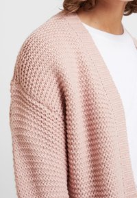 Strickjacke in zartem Rosa, mit strukturiertem Gewebe, offenem Vorderteil und Rippenakzenten entlang des Kragens und der Kanten.