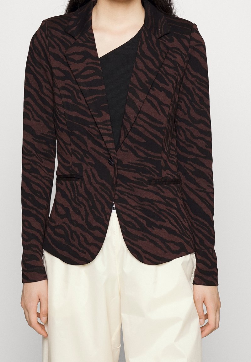Femme portant un blazer cintré à imprimé zèbre noir et marron foncé sur un haut noir et un pantalon blanc ample, debout devant un fond uni.