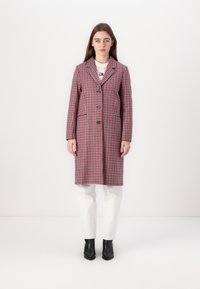Geruite jas in rood, blauw en witte stof, lang met een geknipt kraag, sluiting met één knoop, zijzakken, gecombineerd met witte jeans en zwarte laarzen.