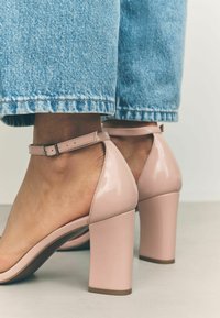 Sandali alti in pelle sintetica rosa con tacco a blocco e cinturino alla caviglia. Pelle esposta sul piede, abbinati a jeans in denim azzurro a gamba larga.