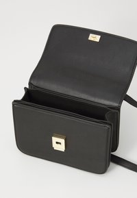 Borsa a mano in pelle nera con una chiusura quadrata dorata. Presenta un unico scomparto e una texture liscia, progettata con un profilo piatto e una tracolla regolabile.