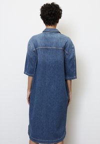 Sukienka koszulowa z denimu w średnim odcieniu niebieskiego. Posiada krótkie rękawy, spiczasty kołnierzyk oraz luźny, sięgający kolan krój z widocznymi detalami szycia.