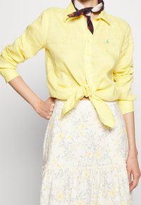 Keltainen pellava nappikauluspaita, jonka hihat on kääritty, vyötärölle solmittuna. Yhdistetty vaalean kukkakuosisen hameen kanssa, jossa on keltaisia ja harmaita yksityiskohtia.