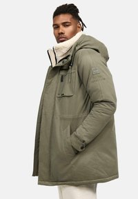 Parka verde con un suave y esponjoso cuello, cierres con cremallera y botones, y dos bolsillos frontales con un logotipo bordado en la manga.