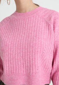 Femme portant un pull côtelé rose doux avec un col montant, un pantalon foncé visible et une petite boucle d'oreille en or.