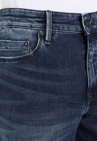 Tmavě modré džínové kalhoty s klasickým designem pěti kapes, vybavené knoflíkovou légou, měděnými nýty a jemným vyblednutím látky.