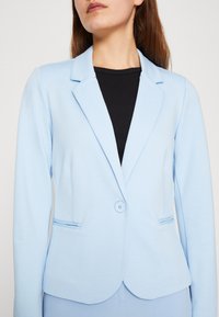 Jasnoniebieska marynarka z klapami, zapinana na jeden guzik, z bocznymi kieszeniami. Wykonana z gładkiego materiału o delikatnej fakturze.