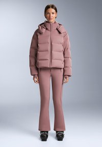 Smeđa prozirna jakna s krojenim dizajnom, s visokim izdignutim ovratnikom i elastičnim manžetama. Uparena sa ružičastim uskim hlačama i crnim skijaškim čizmama.