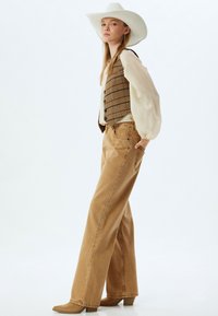 Chaleco marrón a cuadros sobre una blusa color crema, combinado con pantalones de pierna ancha de un marrón claro de cintura alta y botines de tacón beige. Sombrero de vaquero blanco.