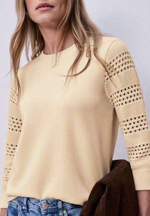 Donna che indossa un maglione beige con maniche lavorate a maglia e con motivi, infilato in jeans di denim blu, che tiene in mano una giacca piegata in camoscio marrone.