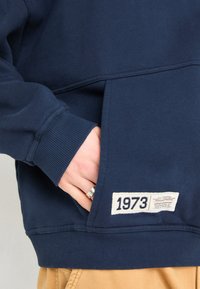 Námořnická modrá mikina s žebrovanými manžetami, která má kapsu a štítek s rokem "1973". Materiál vypadá měkce a má strukturovaný povrch.