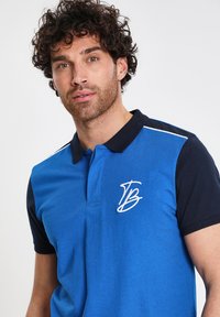 Blå polo skjorta med marinblå ärmar, prydd med en vit broderad logo och krage. Slät textur med klassisk passform och korta ärmar.