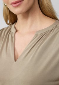 Blouse beige avec un décolleté en V et des détails froncés aux épaules, en tissu lisse. Légère brillance et texture douce.