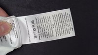 Étiquette d'entretien des vêtements indiquant les instructions de lavage, la composition du tissu 100 % coton et un texte multilingue sur fond noir.