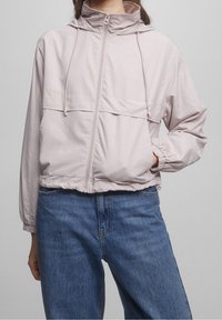 Light pink zip-up jacket with a hood, elastic cuffs and hem, featuring a front pocket and a smooth, lightweight material. Paired with blue jeans.