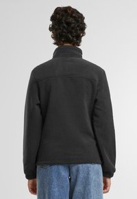 Svart fleece-genser med høy krage, lange ermer og en teksturert overflate. Har en sydd søm over ryggen. Kombinert med blå jeans.