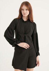 Schwarzes Oberteil mit gerüschtem Kragen und Reißverschluss, ausgestattet mit langen Puffärmeln mit plissierter Textur, kombiniert mit passenden schwarzen Shorts.