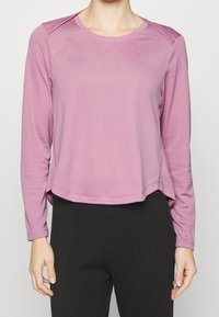 Långärmad topp i mjuk, texturerad rosa tyg med rund halsringning och något kortad fåll. Har minimala sömmar och subtila axelaccenter.