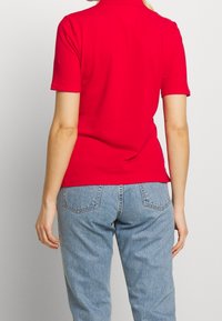 Chemise polo rouge à manches courtes, en tissu coton lisse, avec un col classique et une coupe ajustée. Portée avec un jean en denim bleu clair.
