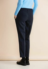 Marinblå byxor med en avslappnad passform, elastisk midja, två bakfickor och nedhasade anklar, i kombination med svarta skor och strumpor.