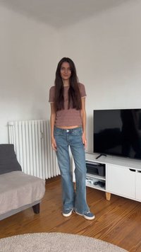 Raudona languota trumpų rankovių palaidinė, aukšto liemens mėlyni džinsai ir mėlynos bateliai su baltais padais. Stovi kambaryje su televizoriumi ir sofa.