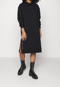 Nainen, joka on pukeutunut mustaan pitkähihaiseen hupparimekkoon, jossa on sivuhalkiot, ja mustiin paksupohjaisiin polven ylle ulottuviin saappaisiin, seisoo yksinkertaisella valkoisella taustalla.