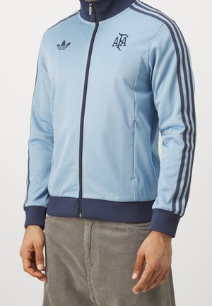 Mann trägt hellblaue Trainingsjacke mit Reißverschluss, marineblaue Streifen an den Ärmeln, navy Kragen und Bündchen und Logos auf der Brust.