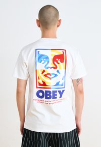 White cotton t-shirt featuring a colorful graphic on the back with faces, patterns, and the word "OBEY" in bold blue. Vertical striped pants.