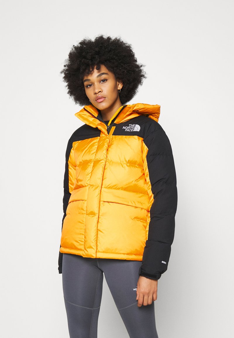 Oranžinė pūkinė striukė su juodomis rankovėmis, turinti gobtuvą, užtrauktuką ir logotipą ant krūtinės. Lygus paviršius, šiltos izoliacijos dizainas.