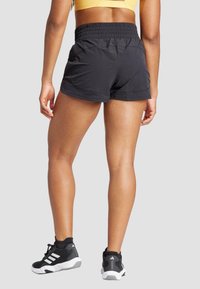 Melni sporta šorti ar elastīgu jostu, gluda virsma un pieguļošs dizains, kopā ar melniem sporta apaviem ar baltām akcentiem.