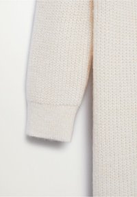 Manche de pull en maille couleur crème avec un poignet côtelé, montrant un tissu en laine texturé sur fond blanc uni.