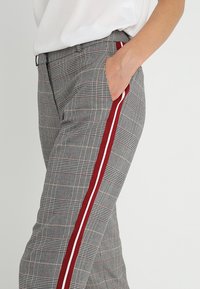 Pantalons à carreaux gris avec des bandes latérales rouges et blanches, présentant une coupe slim et des poches latérales. Le matériau semble texturé et léger.