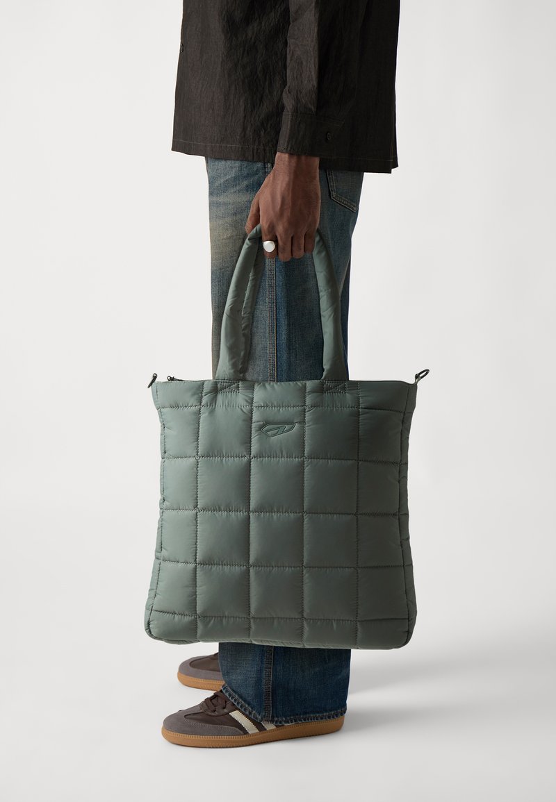 Sac cabas matelassé de couleur vert atténué, fabriqué en tissu rembourré. Il est doté de deux poignées supérieures et d'un logo, avec une forme plate et rectangulaire.