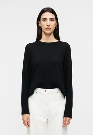 Femme aux longs cheveux noirs portant des boucles d'oreilles créoles en or, un pull noir à manches longues et un pantalon blanc taille haute sur un fond uni.