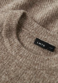 Beżowy sweter o fakturze z okrągłym dekoltem. Metka z tyłu głosi "LMTD" na czarnym tle. Miękki, puszysty materiał o ciepłym wyglądzie.