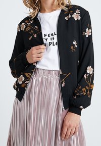 Giubbotto bomber nero con ricami floreali in beige e rosa. Materiale leggero, chiusura con zip e polsini a costine. Indossato sopra una t-shirt grafica e una gonna plissè.