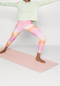 Persona che indossa leggings a gradiente pastello e una maglietta leggera a maniche lunghe, in piedi scalza in una posizione di yoga con gambe divaricate su un tappetino rosa.