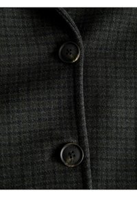 Črn volnen plašč s houndstooth vzorcem, ki se ponaša z gladko teksturo in okroglo, temno gumbom s štirimi luknjami.