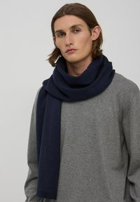 Granatowy dzianinowy szalik luźno opada wokół szyi na tle szarego swetra, z teksturowanym wykończeniem i prążkowanymi końcami.