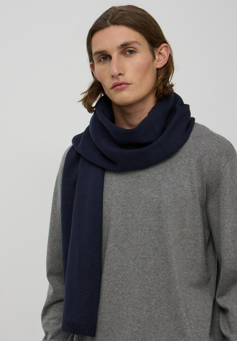 Granatowy dzianinowy szalik luźno opada wokół szyi na tle szarego swetra, z teksturowanym wykończeniem i prążkowanymi końcami.