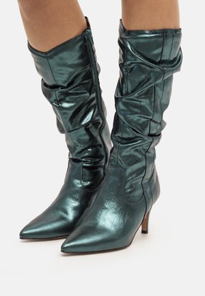 Botas metálicas en color teal hasta la rodilla con punta afilada, detalle de fruncido texturizado y cremallera lateral; presentan un tacón bajo y delgado.